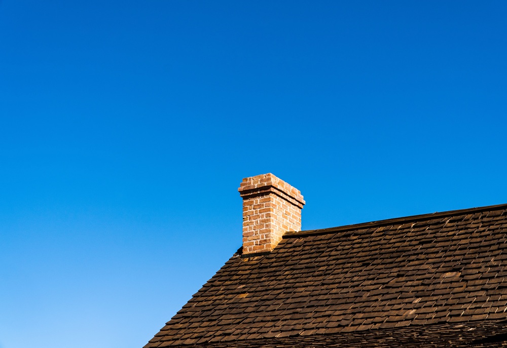 Brick chimney rising above a brown shingled roofline against a clear blue sky